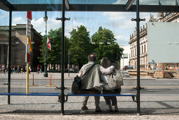 Happy, loving, hugging pensioners, elderly man and woman, sitting at bus stop in Berlin capital of...