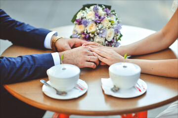 Happy, loving bride and groom, holding hands, sitting in a cafe at a wooden table, drinking coffee, cappuccino from white cups. Wedding photography, portrait of newlyweds.