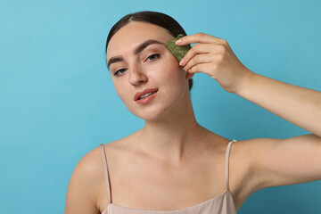 Fototapeta premium Beautiful young woman doing facial massage with gua sha tool on light blue background