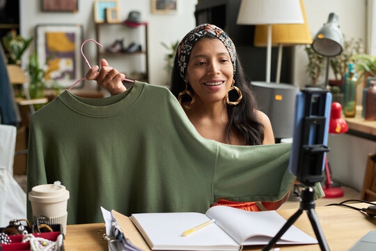 Young smiling woman sitting by desk in front of smartphone and showing new grey long sleeve pullover to online audience