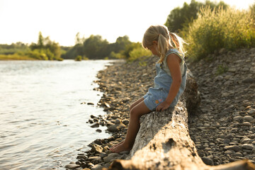 Cute little girl sitting on tree trunk near river. Child enjoying beautiful nature
