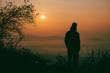 A solitary figure stands softly silhouetted against the warm, glowing hues of an autumn sunrise. Mist rises from the valley as nature awakens to a new day filled with tranquility.