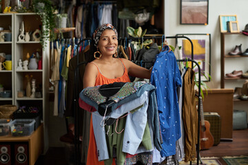 Young cheerful female owner of thrift shop or customer holding stack of casual attire from new...