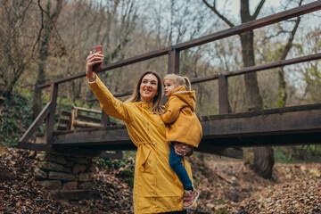 A mother and daughter, both dressed in yellow, pause on a walk through a tranquil forest to take a selfie, with a charming wooden bridge adding to the scenic backdrop.