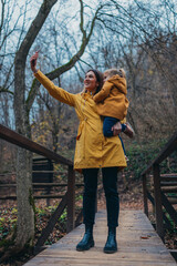 Fototapeta premium A mother and her child are capturing a selfie while standing on a rustic wooden bridge in a serene forest setting, wearing matching yellow jackets.