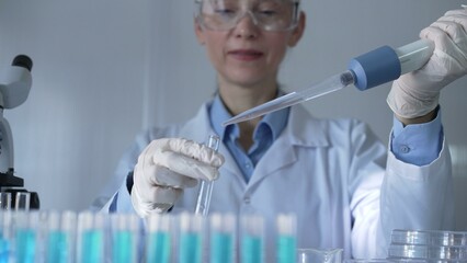 A scientist woman, wearing white gloves and safety glasses, is using a micropipette to drop a...
