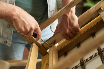 Hands of young unrecognizable male worker of thrift shop fixing screw in bottom part of wooden chair seat with screwdriver