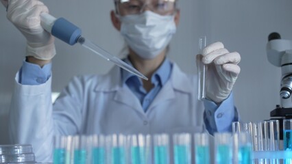 Scientist woman, wearing a lab coat, mask, blue gloves and safety glasses, is using a micropipette...