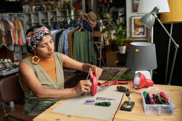 Fototapeta premium Young woman fixing screw in bottom part of electric appliance while sitting by workplace and repairing things in thrift shop