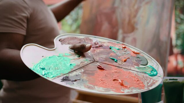 In an art studio, painter mixes multiple watercolor paints on a palette to create artwork on canvas. Skilled man painting with tools and showing imagination, artistic creation. Close up. Camera B.