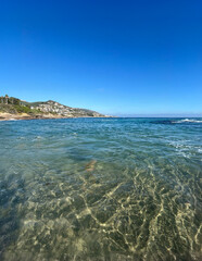 A breathtaking view of crystal-clear ocean water at Laguna Beach, with gently rolling waves and a cloudless blue sky. The sandy seafloor is visible through the transparent water.