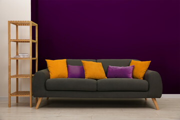 Stylish grey sofa with pillows and shelving unit near purple wall indoors
