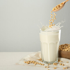 Soy beans falling from wooden spoon into glass of plant milk on white table. Space for text
