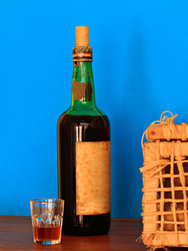 bottle of a very old wine arranged between a glass with a shot and a wicker basket, on a wooden top, against a blue background.
