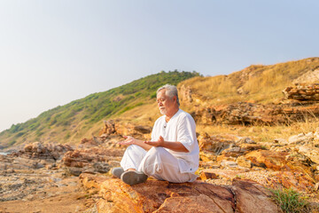 Asian senior man practicing meditation with ocean nature on rocky coastal hill at summer sunset....