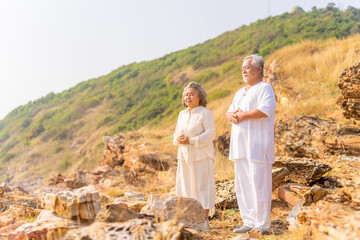 Happy Asian family senior couple practicing yoga and meditation on rocky coastal hill at sunset. Healthy elderly people enjoy wellness and outdoor lifestyle. Mental health care and motivation concept.