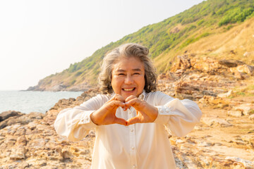 Happy Asian senior woman showing hands heart shape during walking at tropical beach at summer sunset. Retired elderly woman enjoy outdoor lifestyle travel nature ocean on holiday vacation.