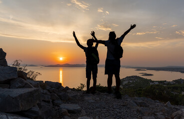 Fototapeta premium Silhouettes of two hikers celebrating at sunset on a rocky mountain with a panoramic view of the sea and distant islands. Concept of outdoor adventure, bonding in nature, and sunset hikes. High