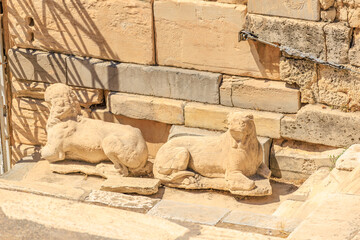 Two statues of lions are laying on the ground in front of a wall, Acropolis in Athens