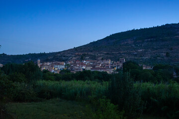 Fototapeta premium Dusk view of a small village on the Iberian Peninsula, seen from a distance in the countryside, with soft street lights beginning to glow. Casas Altas in Rincón de Ademuz, summer 2023