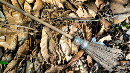 A traditional broom with a wooden handle lies on dry, brown fallen leaves outdoors, surrounded by natural debris. The broom has a bound brush head made from stiff, natural fibers
