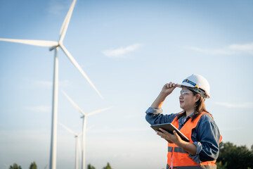 engineer wearing a hard hat and safety vest is looking at a tablet while standing in front of a wind turbine