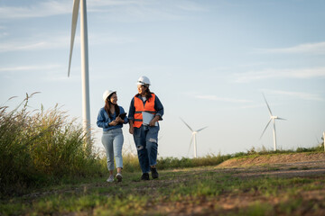 two engineers are walking on a path near a wind farm. One of them is holding a tablet