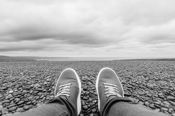 relaxed person wearing sneakers lying on rocky beach looking at cloudy sky, black and white photography perspective shot