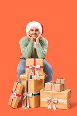Happy young man in Santa hat with gift boxes sitting on chair against orange background