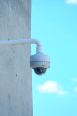 A surveillance camera is mounted securely on a solid concrete wall against a bright blue sky