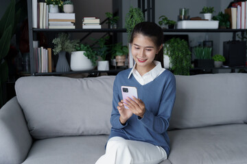 Obraz premium Asian woman sits on sofa using smartphone check social media in living room
