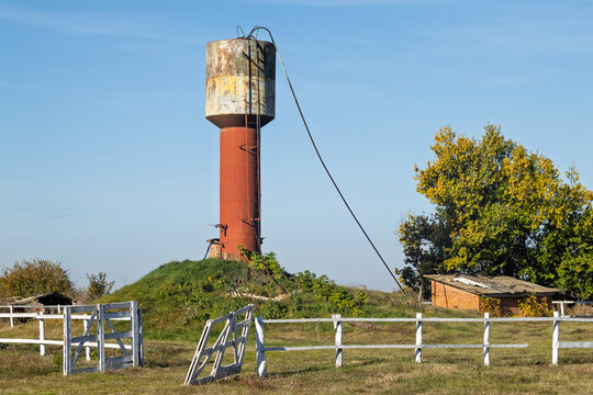 Old water tower in the countryside. A rusty, outdated water tower stands in an open field with a damaged fence and a small brick shed next to it. The scene evokes rural neglect and rural charm.
