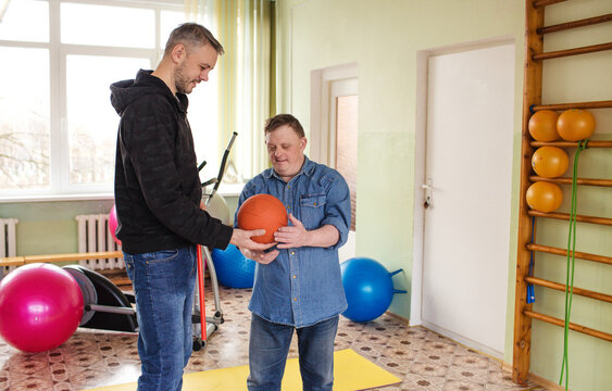 A man with Down syndrome is working out in a gymnasium with his trainer. They are using a medicine ball