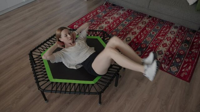 Woman engaging in a workout on a small rebounder trampoline at home, performing abdominal exercises on a colorful rug.