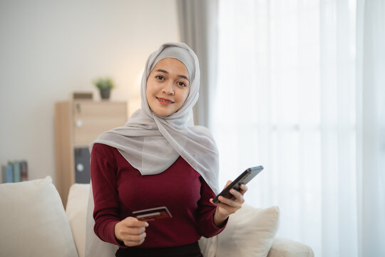 Muslim islam woman with hijab wearing a grey scarf and a red shirt is holding a credit card and a cell phone