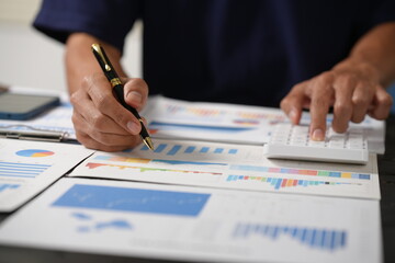 A focused businessman sits at his office desk, analyzing a paper chart while working on his laptop. He examines financial and marketing reports, concentrating on data for strategic decision-making.