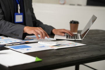 A focused businessman sits at his office desk, analyzing a paper chart while working on his laptop....