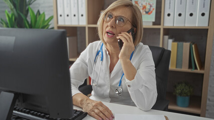 Mature female doctor with blonde hair talking on the phone and working on a computer in a hospital...