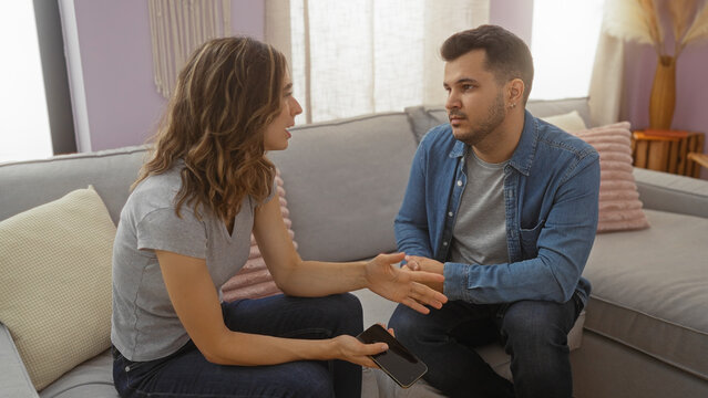 Fototapeta Woman talking with man in living room while holding phone, both sitting on couch, engaging in serious conversation, showcasing indoor relationship dynamics in a home setting