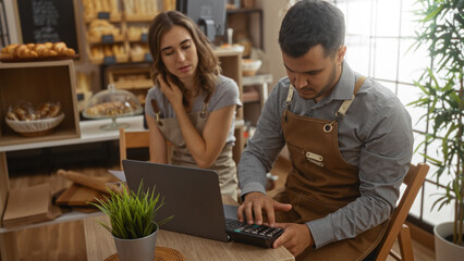 Man and woman in aprons working together on a laptop in a bakery, surrounded by breads and pastries, focusing on calculations and business.