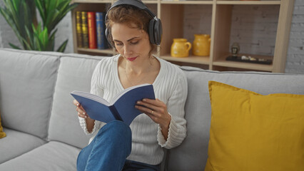 A relaxed caucasian woman enjoys reading a book while listening to music with headphones in a cozy living room.