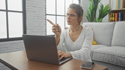 Obraz premium A concerned young woman points at a laptop screen while sitting in a modern living room, indicating an issue or question.