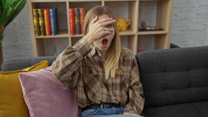 Obraz premium Surprised young caucasian woman with blonde hair covering face with hand in a modern living room.