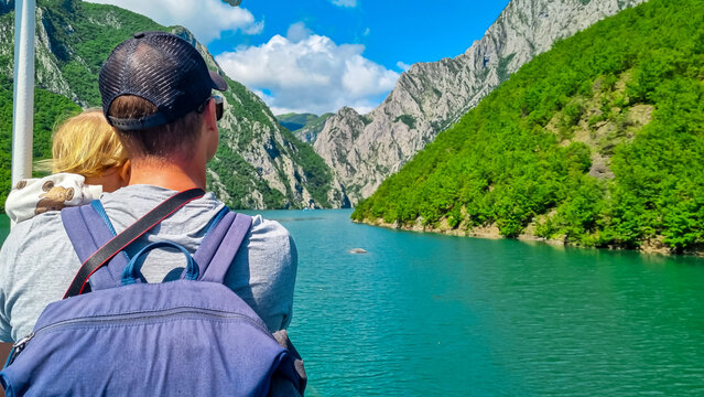 Father holding baby on scenic ferry boat tour on Lake Koman reservoir on Drin river surrounded by forested steep hills, cliffs and mountain ranges of Albanian Alps. Panoramic view of Albanian Fjords