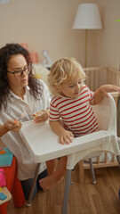 Woman feeding young boy in highchair with toys around in cozy nursery room while creating cherished family moment indoors.