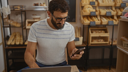 Young man with beard using smartphone in a bakery cafe with shelves of bread and pastries in the background, creating a cozy indoor environment.