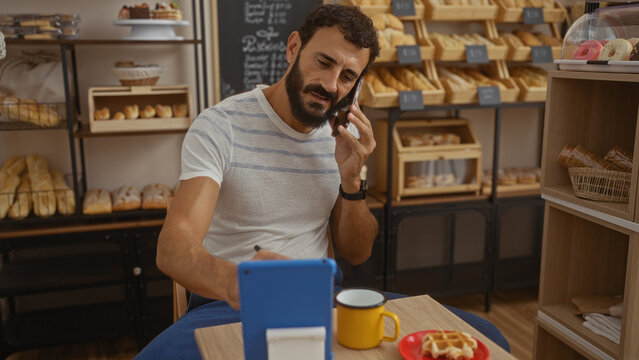 Young man with beard talking on phone while working on tablet in cozy bakery with bread shelves - Powered by Adobe