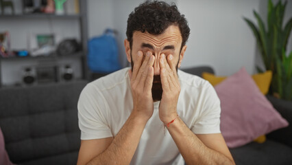 Tired man covering eyes with hands in cozy living room, showing stress or headache.