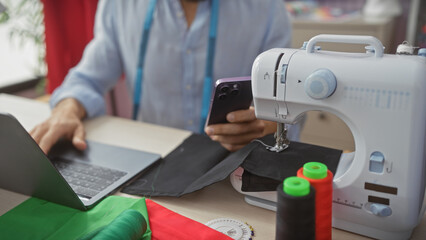 Fototapeta premium Man using laptop and smartphone in a tailor shop amidst sewing materials and a sewing machine