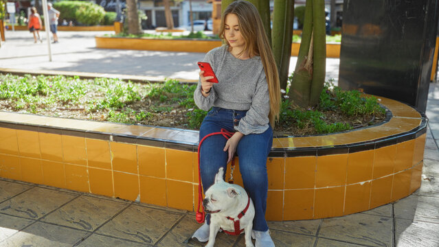 A young, beautiful blonde woman sits outdoors in an urban park, looking at her phone while her pet dog rests beside her.